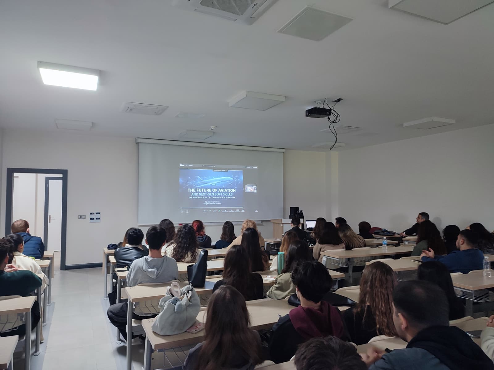 Sınıf ortamında projeksiyon perdesinden Deniz Türkmen'in 'Havacılığın Geleceği ve Yeni Nesil Soft Skill Becerileri' konulu çevrimiçi sunumunu izleyen öğrenciler.